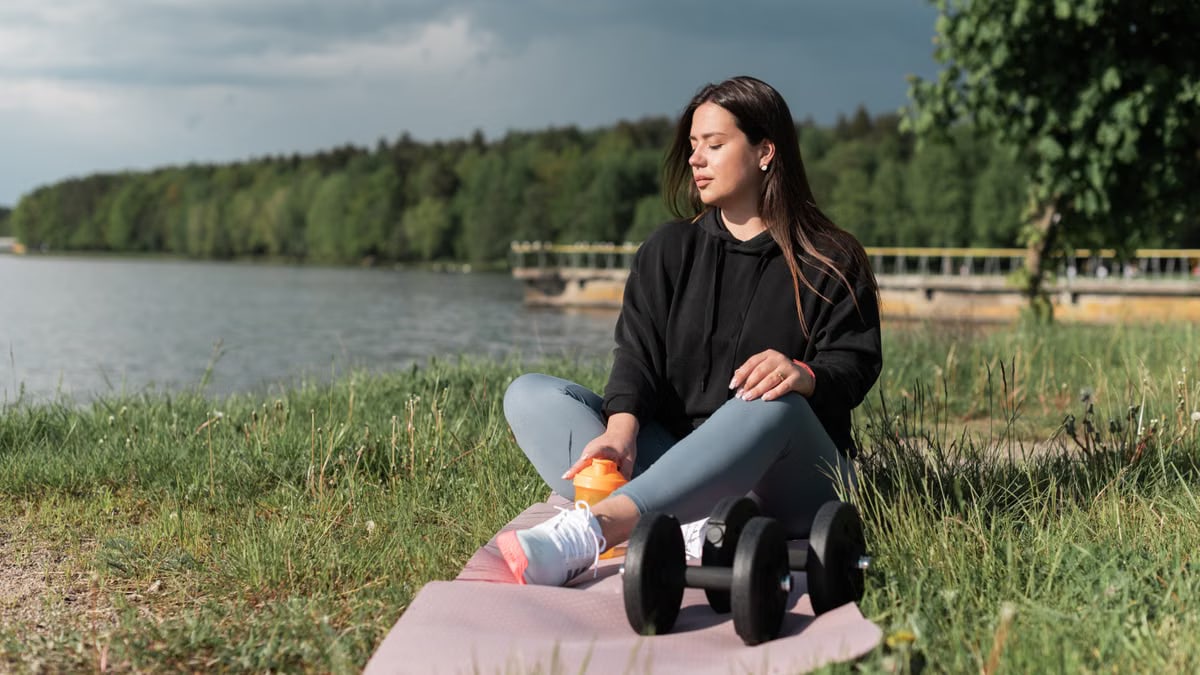 Woman sitting on yoga mat beside a lake with exercise equipment, enjoying a calm outdoor workout.